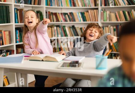 Écolier noir étant intimidé par des camarades de classe, fille et garçon riant et pointant du doigt l'enfant assis dans la bibliothèque ou la salle de classe Banque D'Images