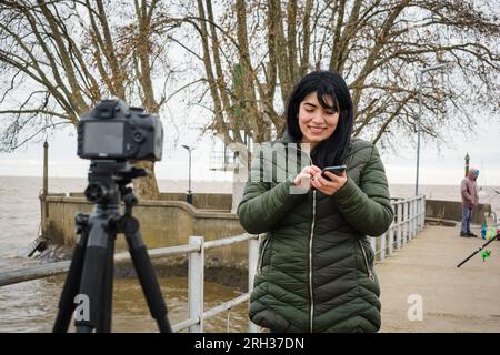 jeune femme vénézuélienne latine créateur de contenu debout avec sa caméra à l'extérieur avec son téléphone dans sa main faisant tutoriel de l'application mobile, co de technologie Banque D'Images