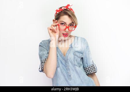 Portrait d'une femme blonde confiante portant une chemise en denim bleu et un bandeau rouge debout regardant la caméra, touchant ses lunettes rouges. Studio intérieur tourné isolé sur fond gris. Banque D'Images