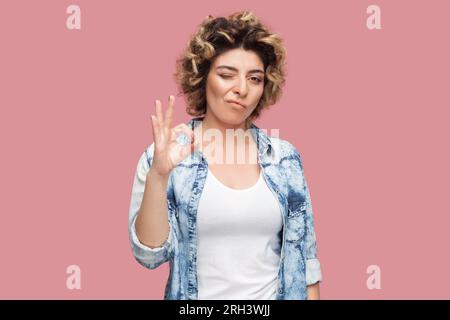 Portrait d'une femme joueuse avec coiffure bouclé portant une chemise bleue debout montrant un geste correct, clin d'œil à la caméra, disant grand travail. Studio intérieur tourné isolé sur fond rose. Banque D'Images