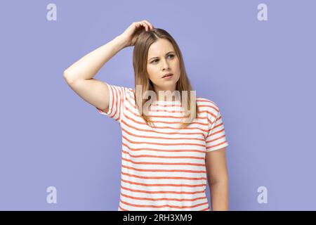 Portrait d'une femme blonde réfléchie intelligente portant un T-shirt rayé pensant à l'idée de démarrage, réfléchissant à la stratégie intelligente, doutant de la décision. Studio intérieur tourné isolé sur fond violet. Banque D'Images