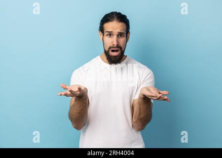Que voulez-vous. Homme confus avec barbe portant un T-shirt blanc debout avec les mains levées et expression indignée surprise, demandant quelle raison. Studio intérieur tourné isolé sur fond bleu. Banque D'Images