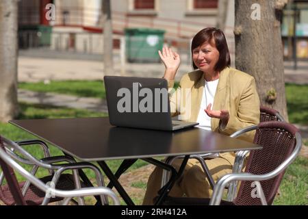 Femme d'affaires adulte élégante, freelance, mentor parle avec un smartphone et parcourt un ordinateur portable au café City Street. Concept de travail à distance, leadership, Banque D'Images