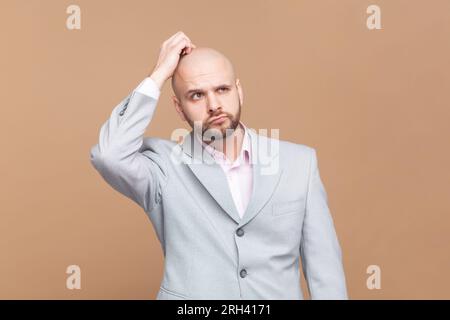 Portrait d'indigné perplexe homme chauve barbu se frotte la tête, essaie de se souvenir des informations nécessaires, regarde dans le mécontentement, portant une veste grise. Studio intérieur tourné isolé sur fond brun. Banque D'Images