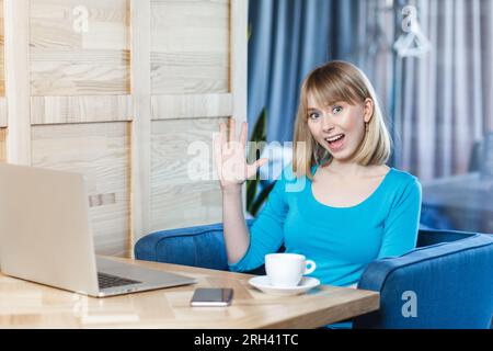 Portrait de jeune femme heureuse amicale avec les cheveux blonds en chemise bleue assis à table, travaillant sur ordinateur portable, rencontrant quelqu'un, agitant la main, disant bonjour. Photo d'intérieur, fond de café. Banque D'Images