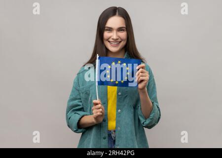 Association européenne. Heureuse femme touristique tenant le drapeau de l'Union européenne, l'immigration et voyageant en Europe, portant une veste de style décontracté. Studio intérieur tourné isolé sur fond gris. Banque D'Images