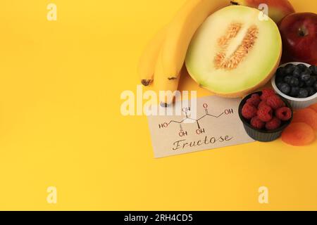 Carte avec structure chimique de fructose, fruits délicieux et baies sur fond jaune, vue au-dessus. Espace pour le texte Banque D'Images