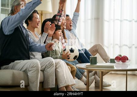 famille asiatique de trois générations assis sur le canapé à la maison célébrant le but et la victoire tout en regardant la retransmission en direct du match de football Banque D'Images