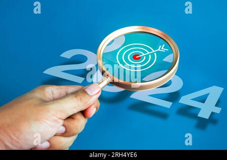 Bonne année 2024 bannière concept. Grande icône de fléchette de cible à l'intérieur de la lentille de loupe tenir à la main tout en regardant le numéro de l'année 2024 sur fond bleu Banque D'Images