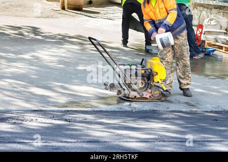 Un travailleur de l'entretien routier verse de l'eau dans le réservoir du compacteur pour compacter ensuite l'asphalte frais à l'aide d'une plaque vibrante à essence. Espace de copie. Banque D'Images