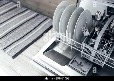 Gros plan de lave-vaisselle automatique en acier inoxydable intégré ouvert à l'intérieur de la cuisine à la maison moderne avec ustensile propre. Assiettes blanches, plats, fourchettes et couteaux en métal. Ménage, ménage, travaux ménagers Banque D'Images