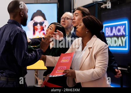 Frénésie les acheteurs de personnes diverses se tenant à l'entrée du magasin deviennent fous pour les offres du Black Friday. Agent de sécurité homme afro-américain gérant le contrôle des foules pendant les ventes saisonnières Banque D'Images