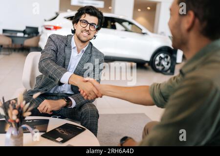 Vue arrière d'un client méconnaissable qui achète une automobile dans un établissement concessionnaire en lui serrant la main du concessionnaire en costume Banque D'Images