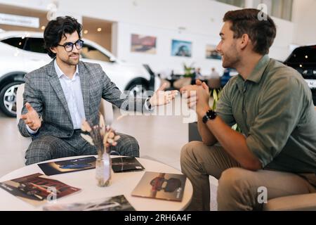 Client masculin heureux parlant des conditions d'achat de voiture avec le directeur portant costume d'affaires assis au bureau dans le concessionnaire automobile. Banque D'Images