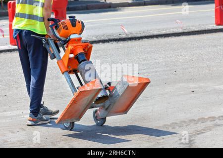 Un travailleur de la route dans un imperméable réfléchissant vert déplace une perceuse à essence sur l'asphalte sur un chantier par une journée ensoleillée. Espace de copie. Banque D'Images