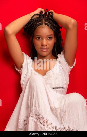 Belle jeune femme noire assise calmement vêtue d'une tenue blanche avec de longues tresses. Isolé sur fond rouge Banque D'Images