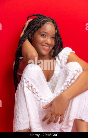 Belle jeune femme noire assise souriante à l'appareil photo. Isolé sur fond rouge Banque D'Images