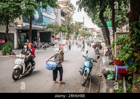 Hanoi, Vietnam, 13 novembre 2022 : une femme âgée portant un panier de légumes navigue en scooter en traversant une rue latérale animée à Han Banque D'Images