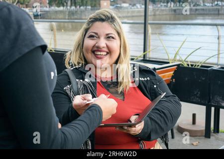jeune femme latine d'origine argentine et de taille supérieure heureuse et satisfaite, elle est assise à l'extérieur du restaurant et donne carte à la serveuse pour la payer Banque D'Images