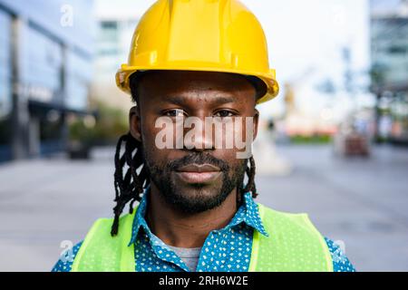 portrait d'un homme d'origine africaine avec barbe et dreadlocks, ouvrier de la construction portant un casque et un gilet réfléchissant, debout à l'extérieur, regardant la caméra Banque D'Images
