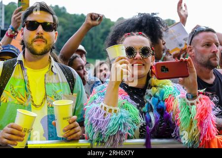 Une jeune femme filmant un numéro sur scène, portant un cardigan tricoté arc-en-ciel multicolore et des lunettes de soleil parmi une foule de gens au Boomtown Fair Festival. Boomtown aka Boomtown Fair est un festival de musique britannique qui se tient chaque année dans le parc national de South Downs, près de Winchester, Hampshire. Depuis 2009, Boomtown a offert un line-up diversifié de groupes, DJs et conférenciers se produisent sur de nombreuses scènes différentes chaque événement annuel est connu sous le nom de Chapitre et se développe sur la ligne de l'histoire de l'année précédente, racontée à travers les décors et les acteurs en direct qui errent sur le site. Banque D'Images