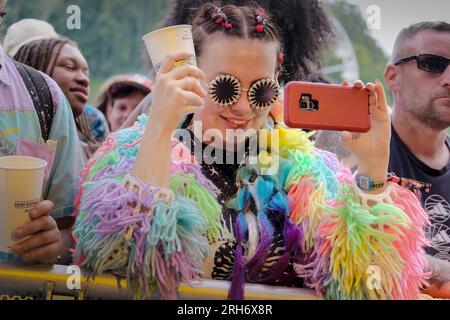 Une jeune femme filmant un numéro sur scène, portant un cardigan tricoté arc-en-ciel multicolore et des lunettes de soleil parmi une foule de gens au Boomtown Fair Festival. Boomtown aka Boomtown Fair est un festival de musique britannique qui se tient chaque année dans le parc national de South Downs, près de Winchester, Hampshire. Depuis 2009, Boomtown a offert un line-up diversifié de groupes, DJs et conférenciers se produisent sur de nombreuses scènes différentes chaque événement annuel est connu sous le nom de Chapitre et se développe sur la ligne de l'histoire de l'année précédente, racontée à travers les décors et les acteurs en direct qui errent sur le site. Banque D'Images