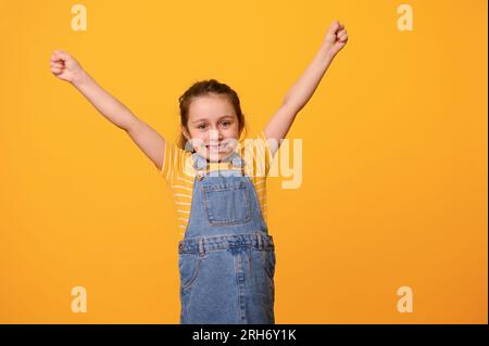 Portrait d'une joyeuse souriante joyeuse petite fille charismatique d'âge préscolaire, portant une robe en denim bleu, serrant les poings et levant les bras, exprimant positiv Banque D'Images