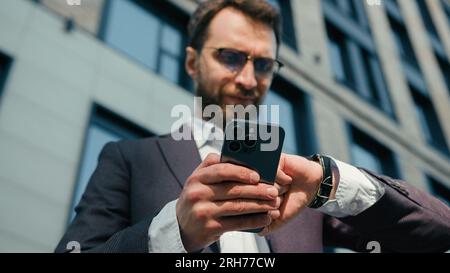 Homme caucasien tenir le téléphone portable à l'extérieur occupé homme d'affaires nerveux vérifier le temps au message texte de montre-bracelet sur smartphone stressé gars affaires Banque D'Images