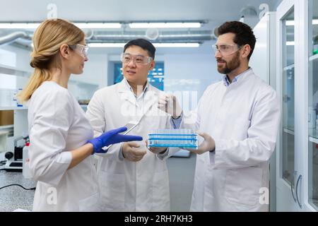 Équipe internationale de médecins, scientifiques, homme et femme debout en laboratoire en blouse blanche, tenant des flacons en verre et discutant de la recherche. Banque D'Images
