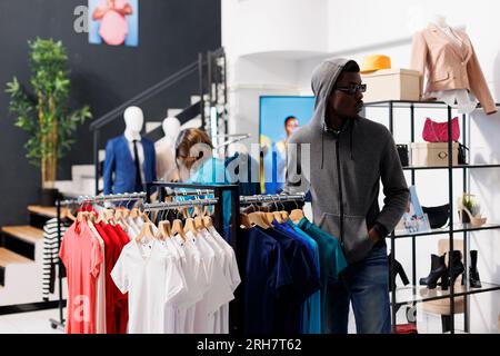 Homme afro-américain avec capuche et lunettes, essayant de voler des marchandises à la mode du magasin de vêtements. Voleur regardant autour de lui pour voir si quelqu'un le regarde tout en volant des vêtements à la mode Banque D'Images