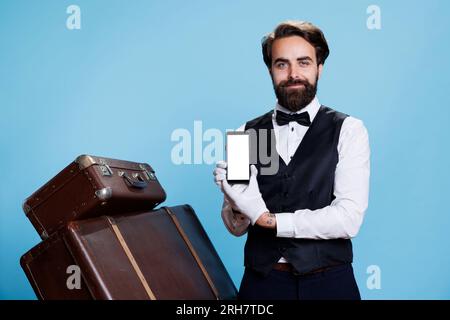 Le portier chic montre l'écran blanc sur le smartphone, tenant le dispositif pendant qu'il est en tenue d'hospitalité professionnelle. Élégant groom déterminé présente un modèle d'espace copie vierge sur l'affichage. Banque D'Images