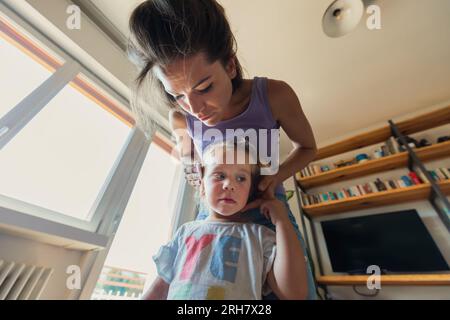 une vue grand angle d'en haut montre une maman fixant les cheveux de sa fille dans un salon lumineux rempli de livres et de multimédia. La croissance est nourrie Banque D'Images