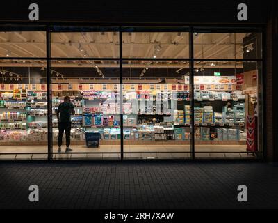 Un regard par une fenêtre dans un supermarché la nuit. L'homme se tient debout avec le téléphone portable. Copenhague, Danemark - 14 août 2023. Banque D'Images