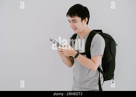 Photo de mise au point sélective d'un jeune homme asiatique portant un T-shirt décontracté vert sauge, une montre intelligente et un sac à dos utilisant un smartphone avec expression de visage souriant Banque D'Images