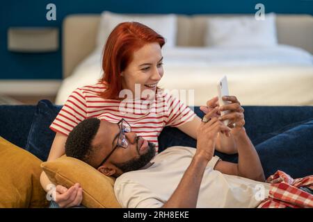 Multi ethnique heureux couple homme et femme regardent le téléphone allongé sur le canapé dans le salon. Banque D'Images