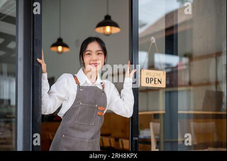 Une jeune femme asiatique joyeuse propriétaire de café ou serveuse dans un tablier se tient à la porte d'entrée de sa boutique. restaurant, boutique, magasin, café Banque D'Images