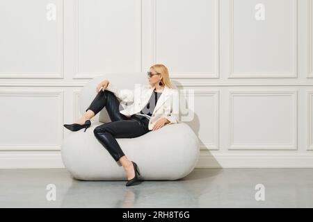 Femme blonde en cuir noir pantalon, chemisier et veste en laine blanche assis dans le profil Banque D'Images