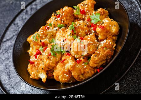 bouquets de chou-fleur d'orange croustillants saupoudrés de graines de sésame, de flocons de piment et de persil dans un bol noir sur une table en béton, gros plan Banque D'Images