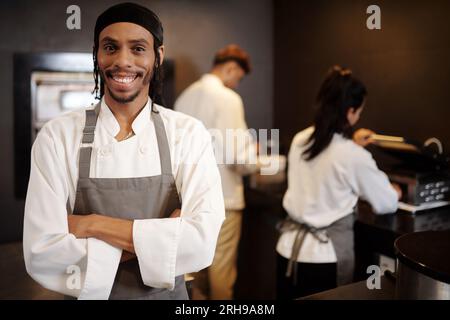 Portrait de restaurant souriant sous-chef en chemise blanche et tablier gris debout dans la cuisine Banque D'Images