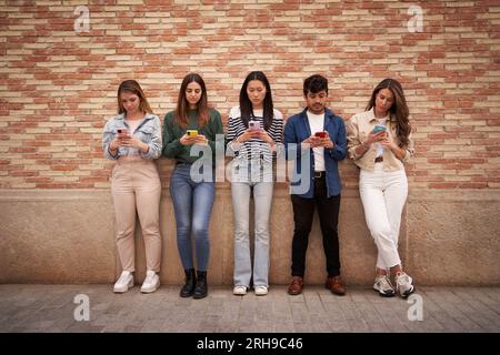 Groupe sérieux multiracial jeune génération z se penchant sur le mur de briques en utilisant des téléphones à l'extérieur. Inquiet. Banque D'Images