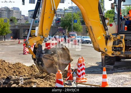 Le godet d'une pelle de construction creuse une tranchée sur la route avec pour toile de fond une rue de la ville. Banque D'Images