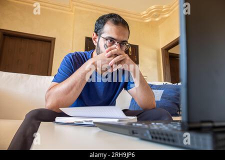 Jeune homme arabe travaillant à la maison avec un ordinateur portable et des papiers sur le bureau Banque D'Images