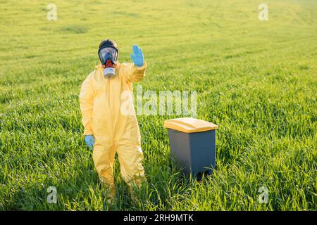 Une femme en costume de protection jaune se tient au milieu d'un champ vert dans un masque à gaz de protection et montre un geste d'appel pour arrêter et sauver le pl Banque D'Images