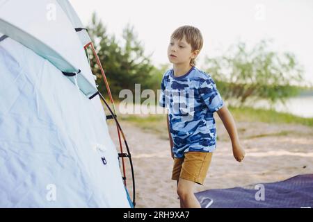 mignon petit garçon caucasien aidant à monter une tente. ombres de coucher de soleil des arbres. Concept de camping familial. Photo de haute qualité Banque D'Images