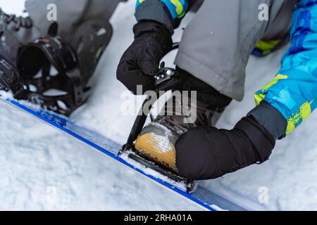 close up vie de petit garçon assis sur la neige mettant ses pieds dans des fixations de snowboard ajustant des sangles. Image avec mise au point sélective. Photo de haute qualité Banque D'Images