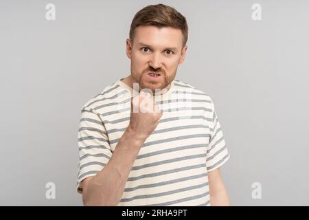 Homme en colère montrant le poing avec grimace mécontente et yeux fous regardant la caméra isolée sur le mur gris Banque D'Images
