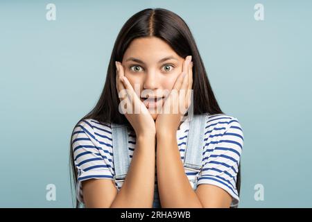 Émerveillé jeune fille heureuse couvrant les joues avec des paumes bouche ouverte isolé sur fond bleu studio Banque D'Images