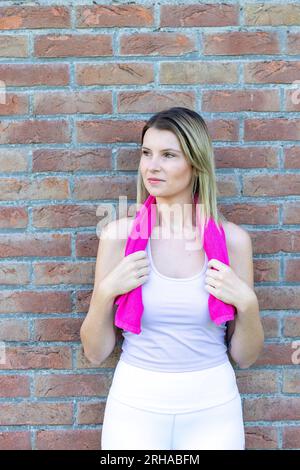 Une femme de fitness radieuse et énergique, vêtue d'un haut sportif et de leggings, prend une pause bien méritée après une séance d'entraînement rigoureuse. Elle sourit avec un sentiment d'accomplissement tout en essuyant la sueur de son front avec une serviette. Cette image incarne les concepts de bien-être, de vitalité et de détermination d'un coureur ou d'un joggeur actif. Femme énergique Fitness : souriante et rafraîchie après l'entraînement. Photo de haute qualité Banque D'Images