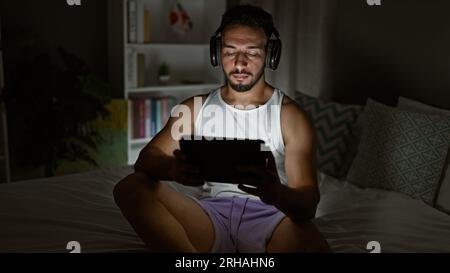 Jeune homme arabe utilisant le touchpad assis sur le lit portant des écouteurs dans la chambre Banque D'Images