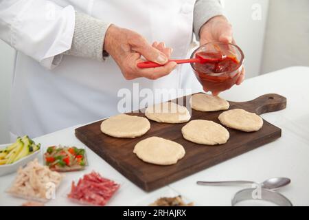 Femme senior ajoutant de la sauce pizza à la pâte étirée. Délicieuse préparation de mini pizzas maison. Banque D'Images